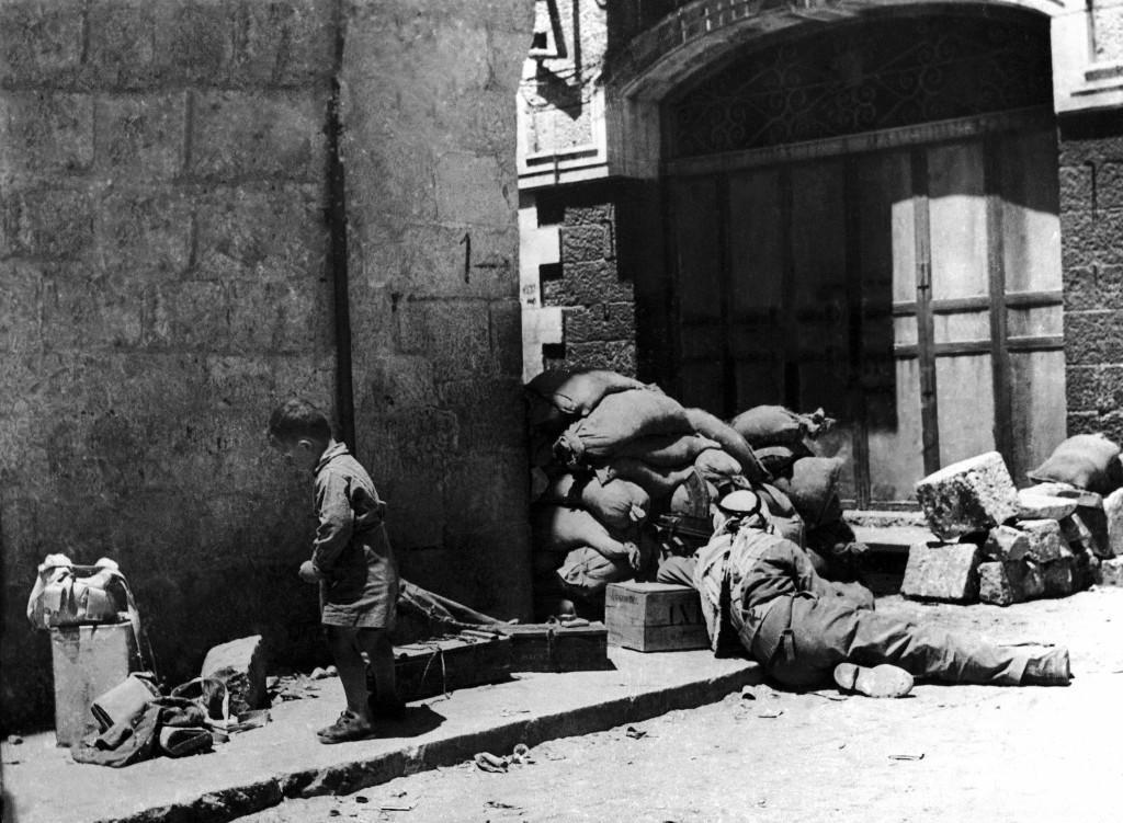 An Arab soldier manning a machine gun, takes cover behind sand bags on a street near the New Gate in the old city of Jerusalem, Palestine on May 29, 1948 during the Jewish Arab conflict while a little boy on the left, unimpressed by the deadly atmosphere plays in the debris covering the pavement. (AP Photo) Ref #: PA.10005843  Date: 29/05/1948