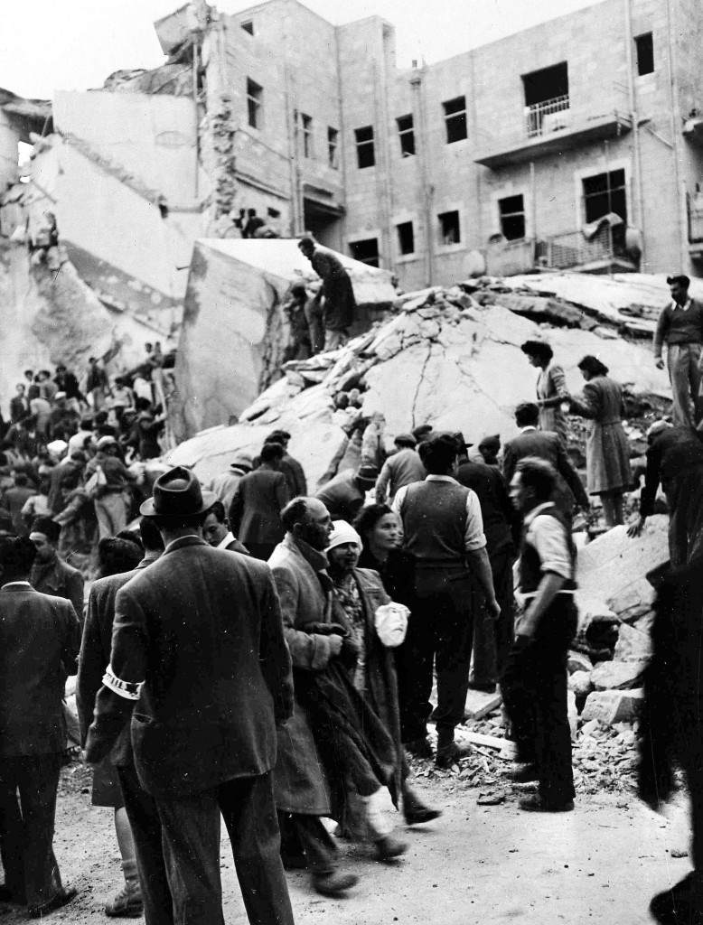 An injured Jewish couple walk past rescuers searching for wounded and dead people in the wreckage of shops on Ben Yehuda Street, Jerusalem, Feb. 22, 1948, after a bomb exploded. The bomb killed 52 Jews and wounded 100 more and levelled buildings on both sides of the street. (AP Photo/Pringle). Ref #: PA.5737217  Date: 22/02/1948