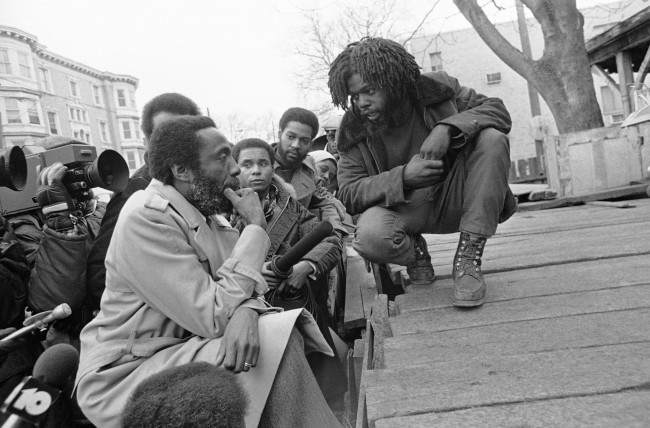Dick Gregory, political satirist and activist, left, talks with man calling himself Phil Africa in front of the MOVE headquarters in Philadelphia's near west side, . Gregory has agreed to help mediate the long stand-off between the city and the radical organization. Date: 10/03/1978