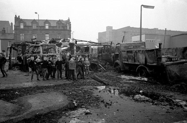 Schoolboys cheer adn chant from a pile of burnt-out busses and lorries in the aftermath of the riots. Date: 09/02/1971