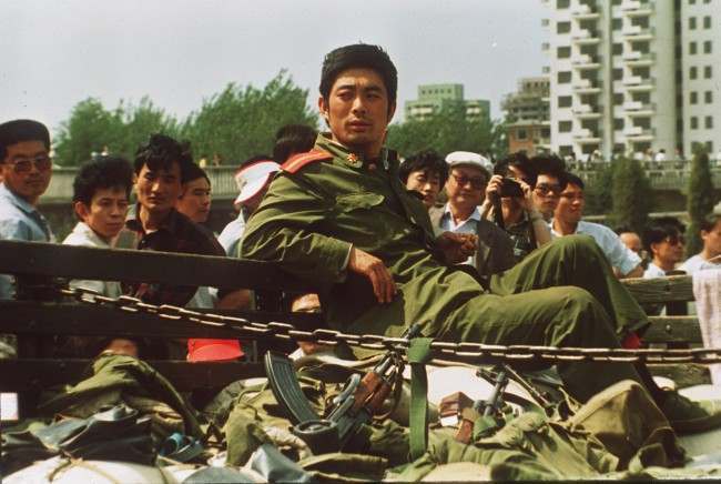 A People's Liberation Army Soldier sits alone with his weapons Sunday after his convoy was stopped by demonstrators seen in the background. (AP Photo/1989)