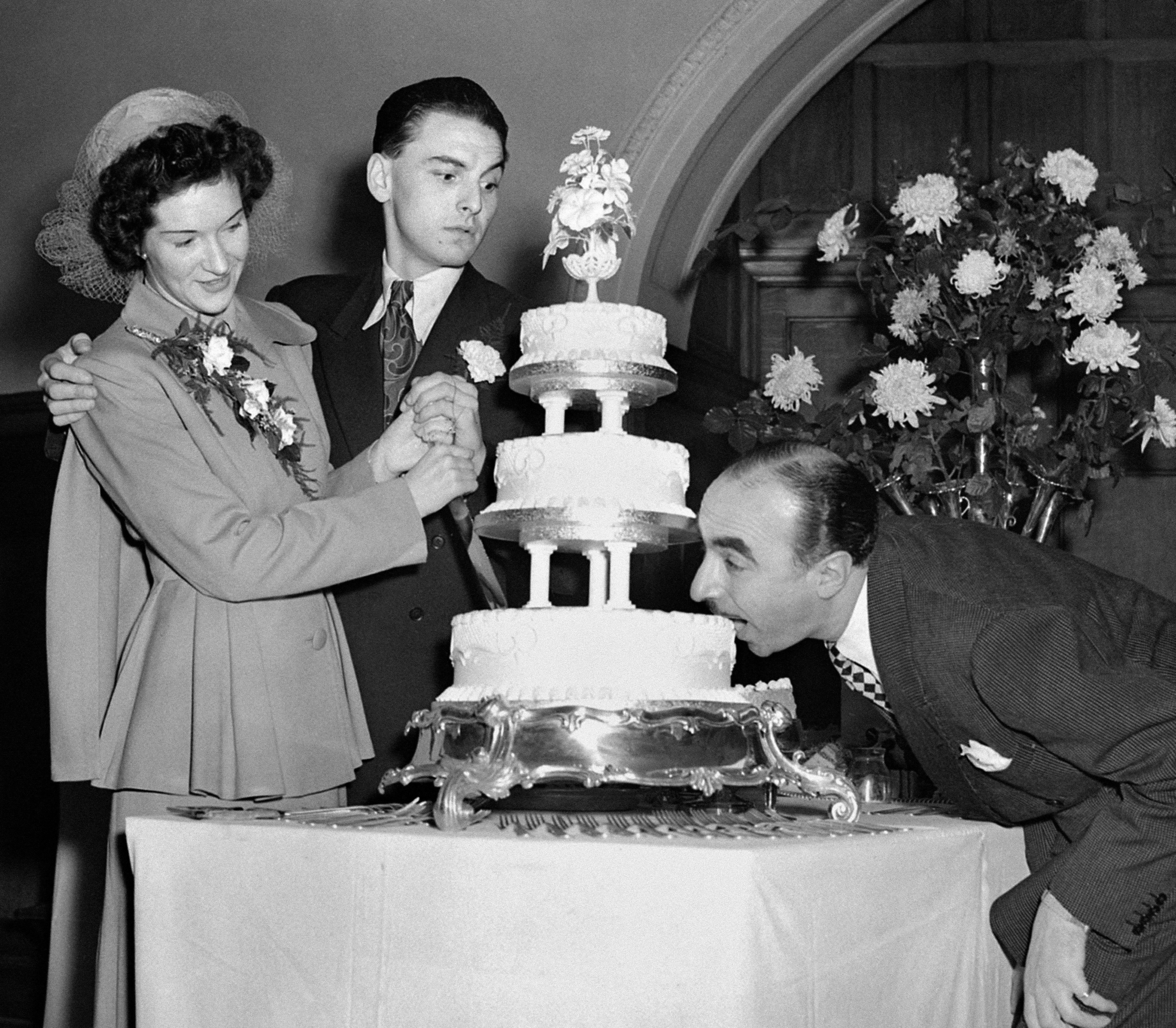 comedian Bob Monkhouse and his bride Elizabeth Thompson, a former nurse from Belfast at their wedding reception at Caxton Hall, London. Harold Berens is taking a bite of their cake as they attempt to cut it.