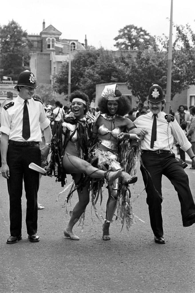 Police constable John Corbett joins an impromptu knees-up with Notting Hill Carnival revellers. PC Corbett, from Harrow Road police station, found his can-can partners on the second day of the August Bank holiday festival. Ref #: PA.7740097  Date: 30/08/1982