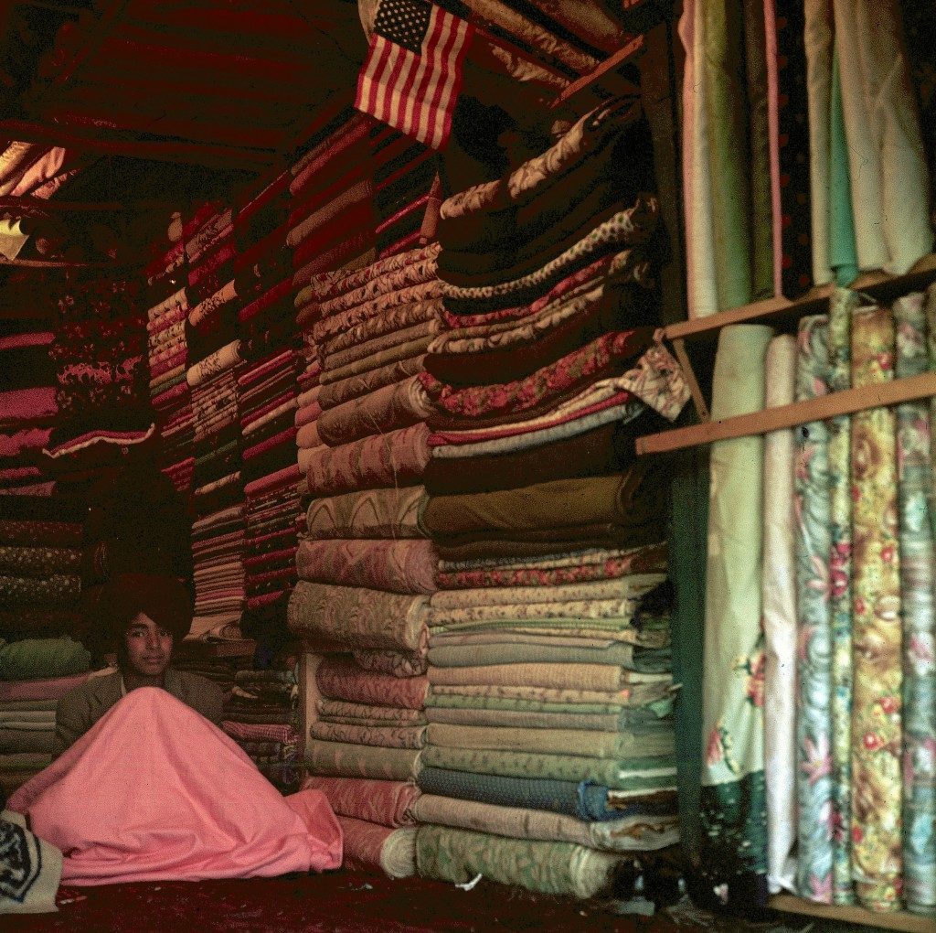 A shopkeeper is shown in a cloth shop where a small American flag is displayed among the rows of folded fabric in the old quarter of Kabul, capital city of Afghanistan - 1960 