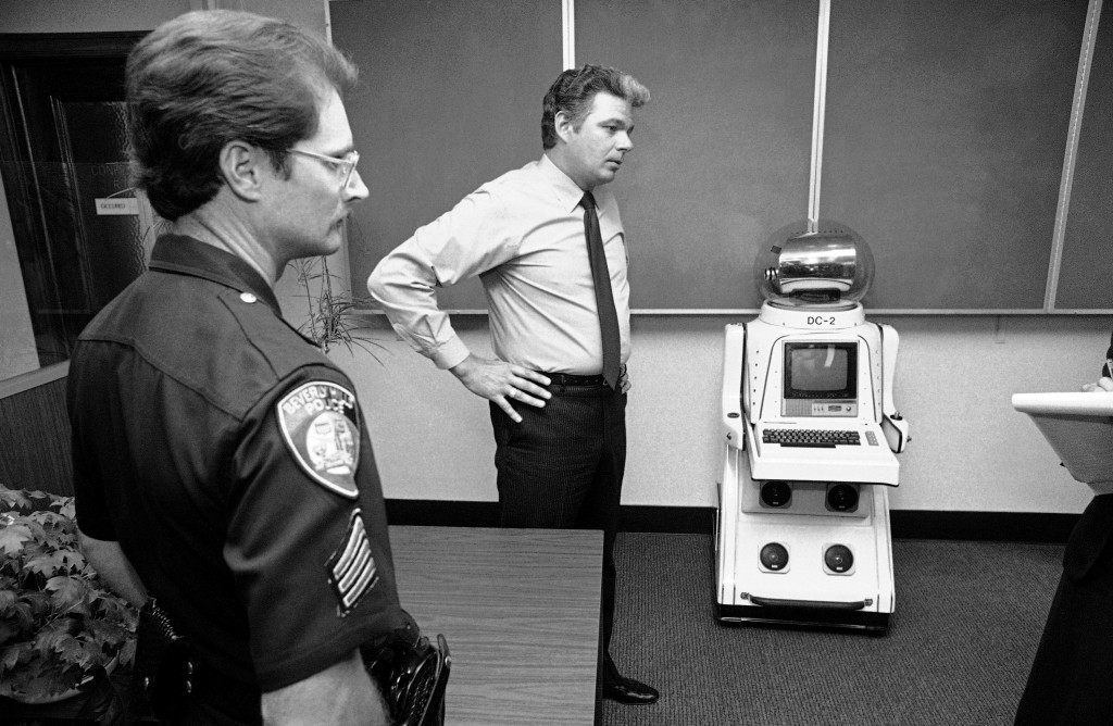Sgt. Tom Van Ansdell of the Beverly Hills, Calif., police department displays a 4-foot robot at police station in Beverly Hills on August 18, 1982. The robot, complete with color television screen and camera, micro computer and two-day communications, was taken into police custody after it was found clanking through Beverly Hills during Tuesday rush hour. Police took the robot into custody when the person, as yet unidentified, operating it by remote control, refused to identify himself to police. (AP Photo/Lennox McLendon)