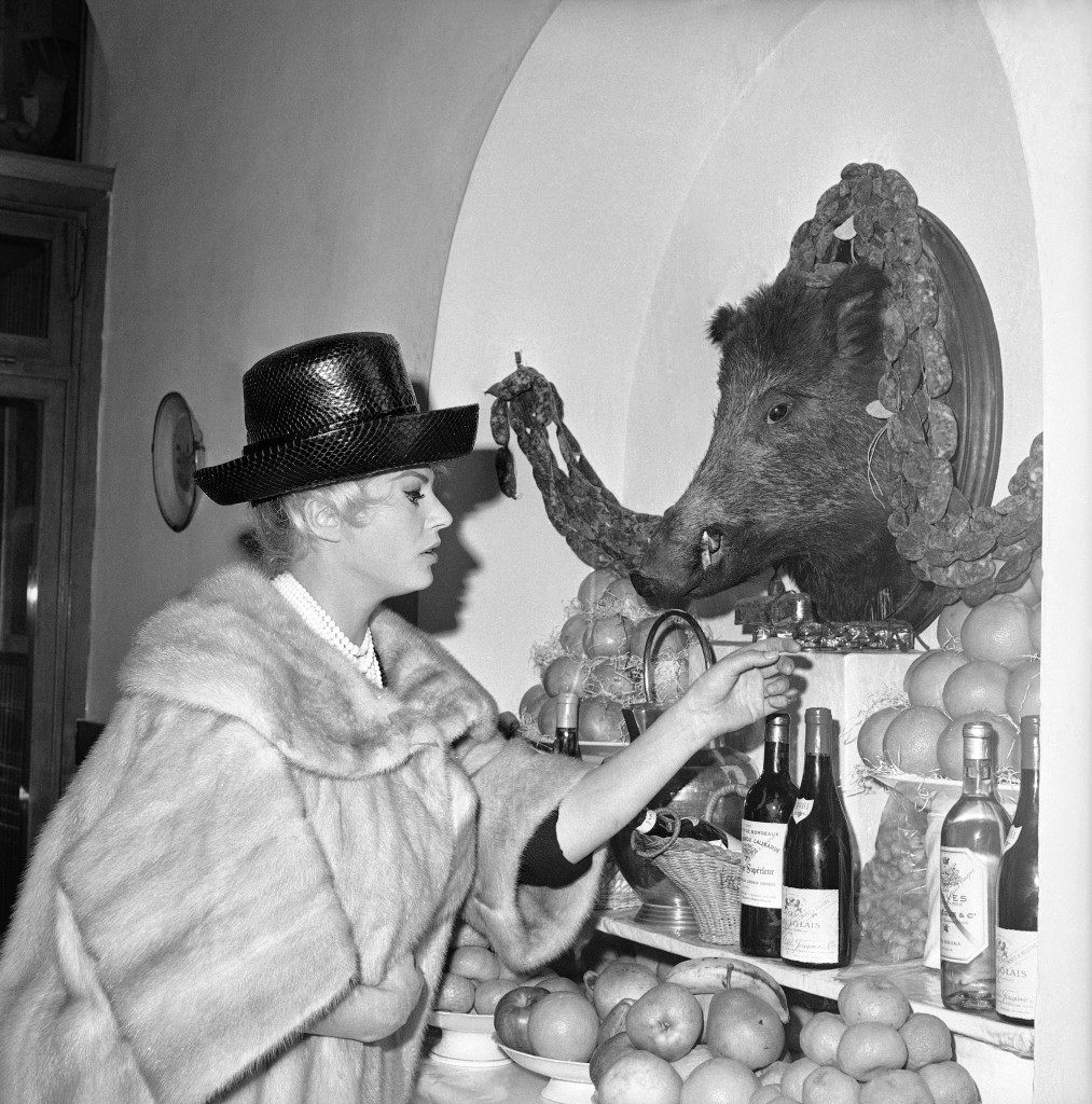 Swedish-born actress Anita Ekberg chooses a bottle of wine for a lunch she and her husband, Austrian-born American actor Frederick "Rik" Van Nutter, are going to have in a fashionable Rome restaurant, Italy on Feb. 26, 1964, famed for its dishes of game (note the head of a boar on the wall, decorated with game-sausages). (AP Photo/Giulio Broglio)