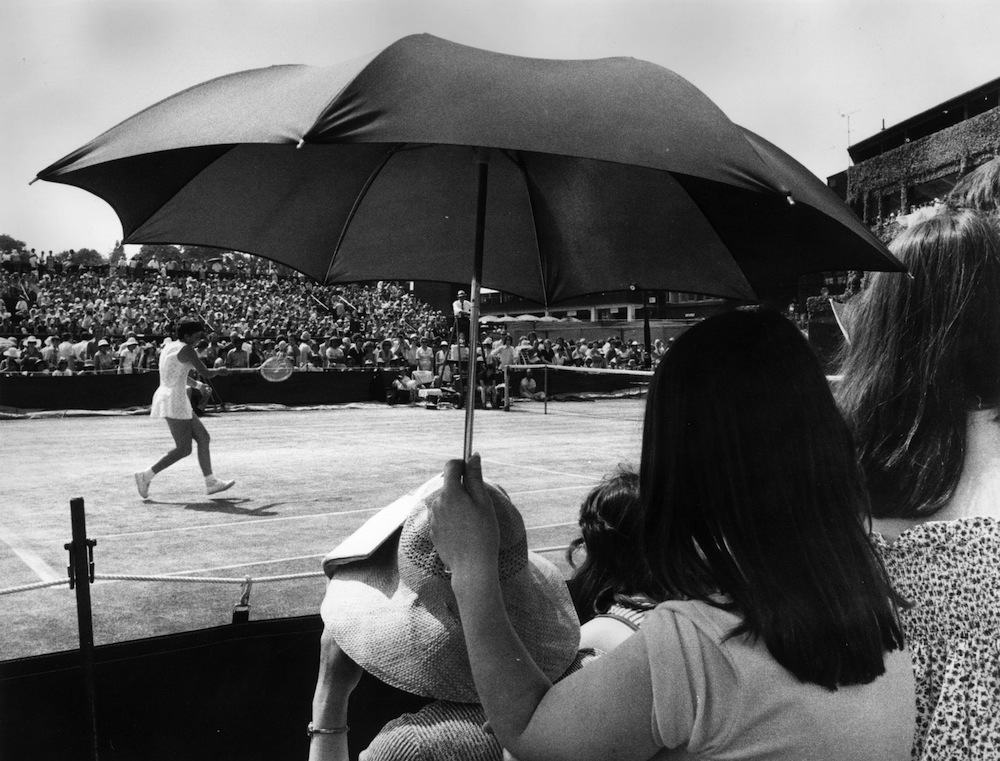 25th June 1976:  A spectator at the Wimbledon Lawn Tennis Championships using an umbrella to shelter from the sun, as Marise Kruger of South Africa plays a match.  (Photo by Roger Jackson/Central Press/Getty Images)
