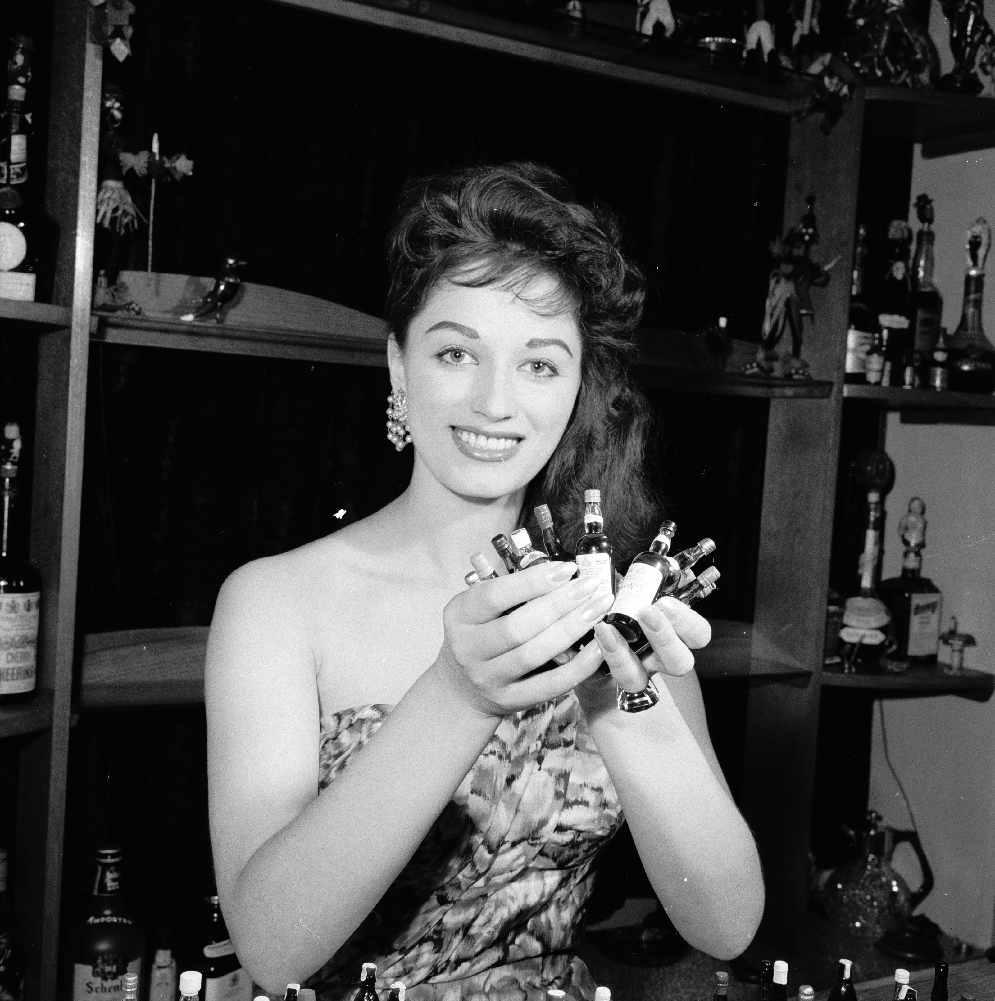 September 1958:  English actress and novelist Jackie Collins at the cocktail bar in her home in London proudly displaying her collection of miniature bottles.  (Photo by Frank Martin/BIPs/Getty Images)