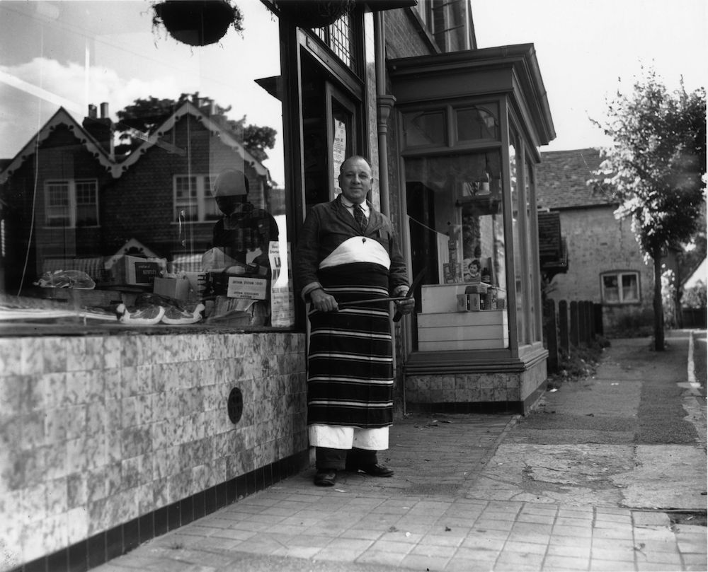 19th August 1953: A village butcher stands outside his shop in Shere, Surrey. Reflected in his shop window are typical Surrey houses. (Photo by T. Marshall/Topical Press Agency/Getty Images)