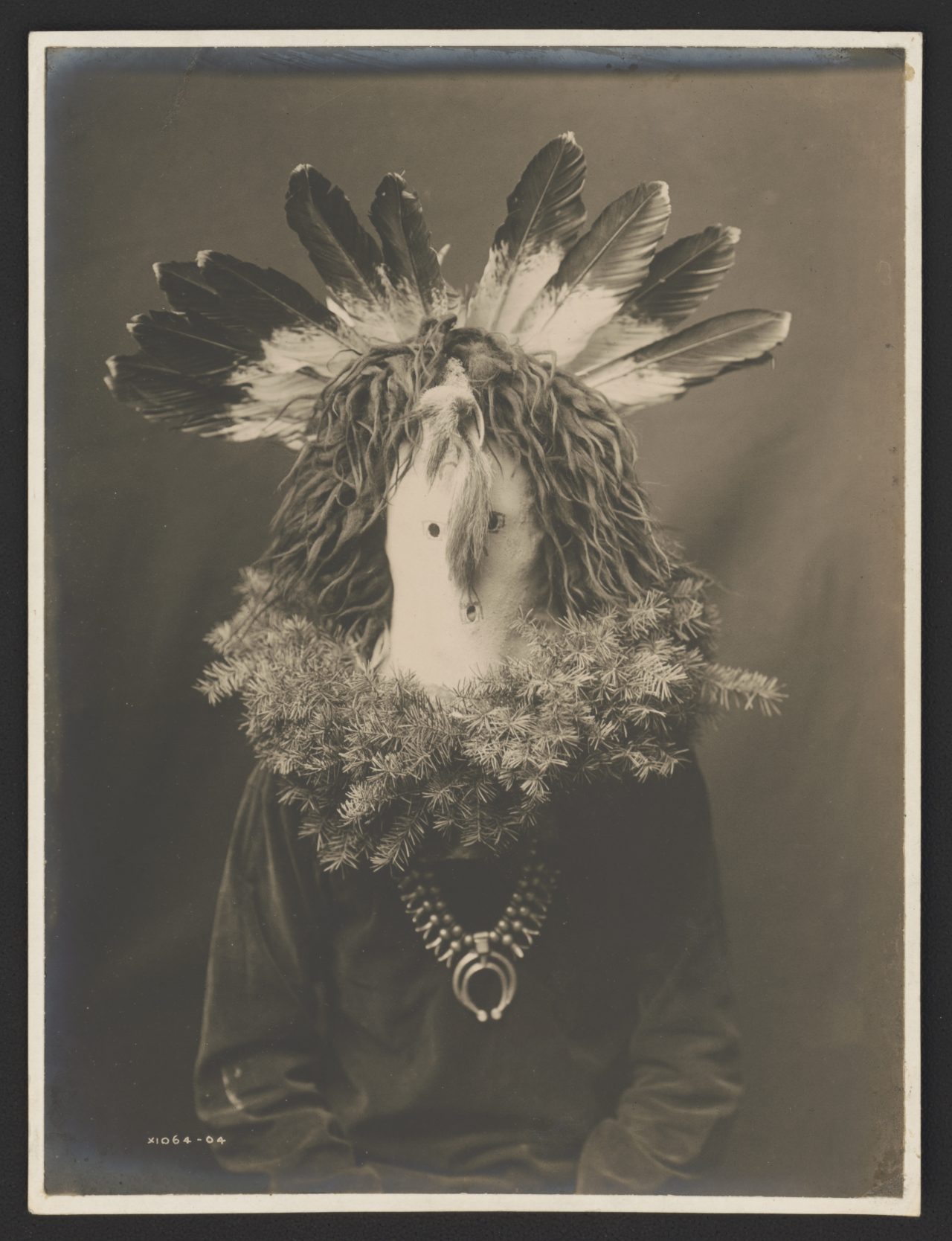 Title Haschogan (House God) - The Yebichai Hunchback Summary Photograph shows a Navajo man, half-length, seated, facing front, wearing a ceremonial mask with feathers and with fir or spruce branches forming a wreath around the shoulders. Contributor Names Curtis, Edward S., 1868-1952, photographer Created / Published [1904]