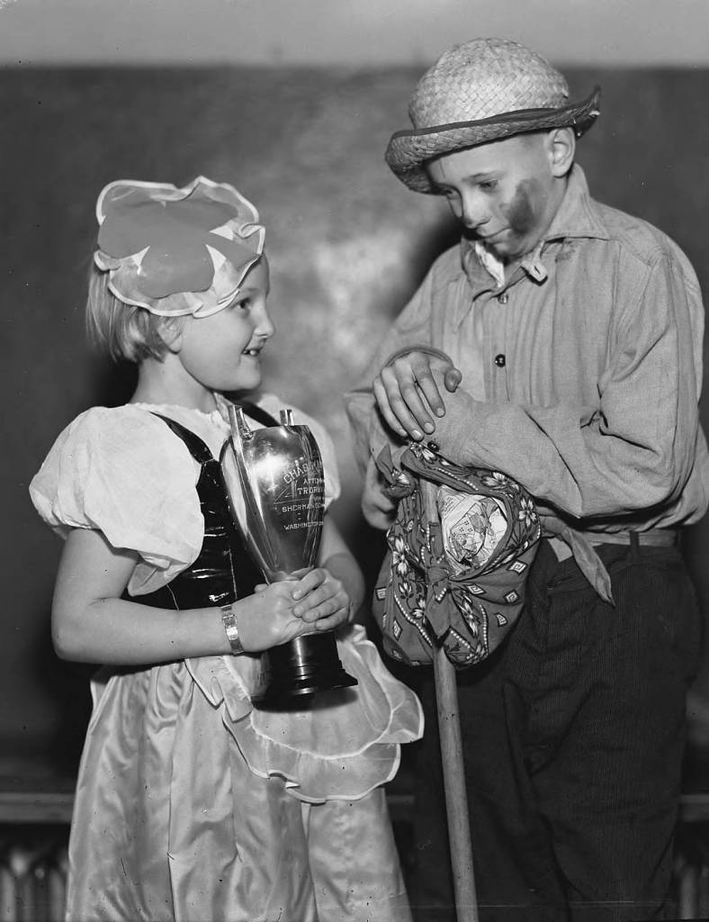 Marilyn Wagnild of Jefferson Elementary School, dressed as "Bo-Peep" and "hobo" Edward Miller of Point Defiance School were just two of the several thousand youngsters and parents who took part in the 1935 Halloween party sponsored by the 26th and Proctor Street Business Mens' Club. The celebration began with a costume parade, led by Mayor George A. Smitley and the American Legion drum and bugle corps, beginning at 7:30 p.m. at Proctor and Stevens and ending at Mason school grounds where prizes were awarded and a huge bonfire was held.. The school with the most participants received the trophy being held by Miss Wagnild. (T. Times, 10/31/1935, p. 1)