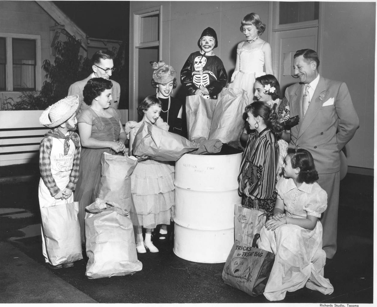 Description Two unidentified men watch as nine unidentified Proctor area "trick-or-treaters" deposit the clothing that they collected for shipment to Korea, during the Halloween weekend, into marked white barrels. In 1953, the Oil Information Committee of Pierce County was working with the Overseas Relief Committee of the Tacoma Council of Churches to collect warm clothes for the needy in Korea. Items needed included blankets, coats and sensible shoes, children's shoes & clothes, baby layettes and knitting wool and sewing thread. Donations could be taken to service stations and deposited in special white barrels marked "Clothing for Korea".