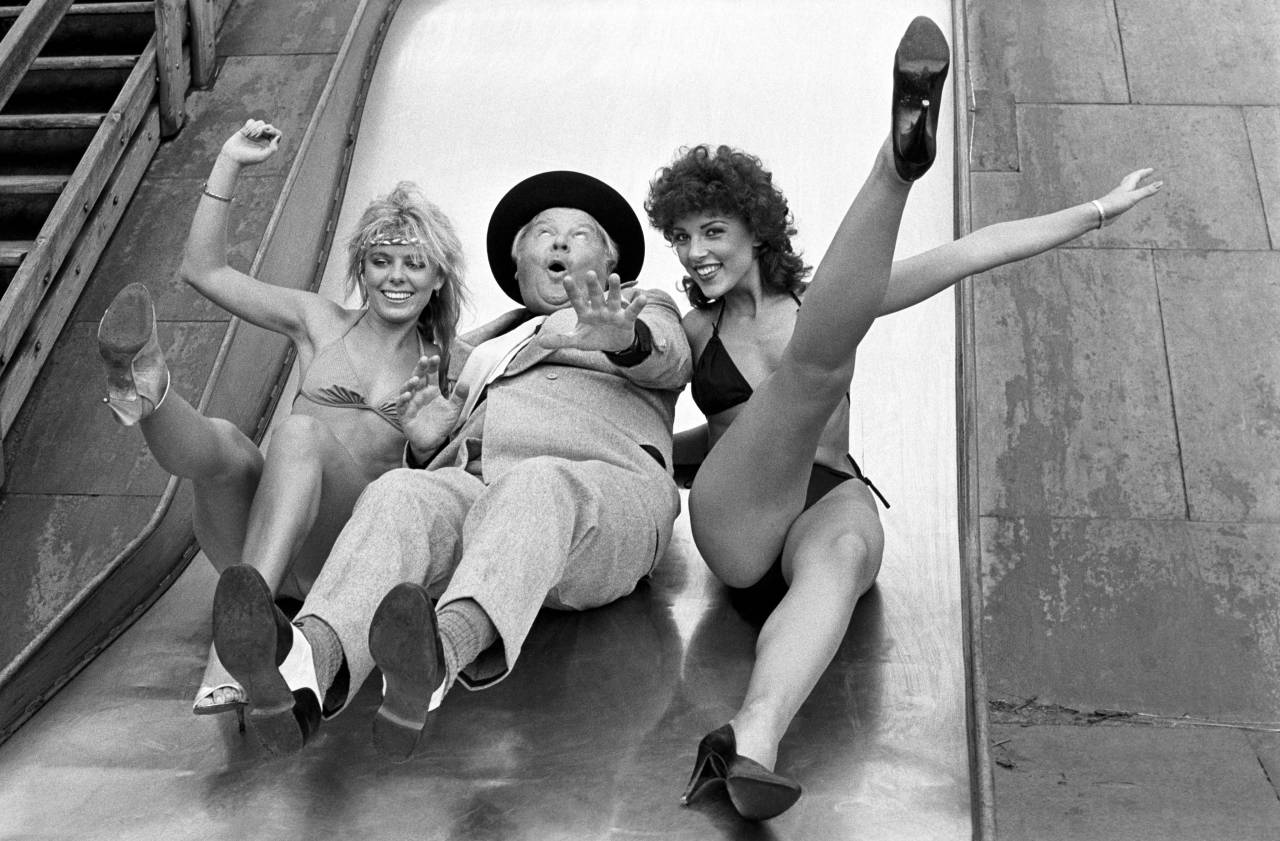 Comedian Benny Hill slides down a slide with two bikini clad women during filming for his latest series (1983).