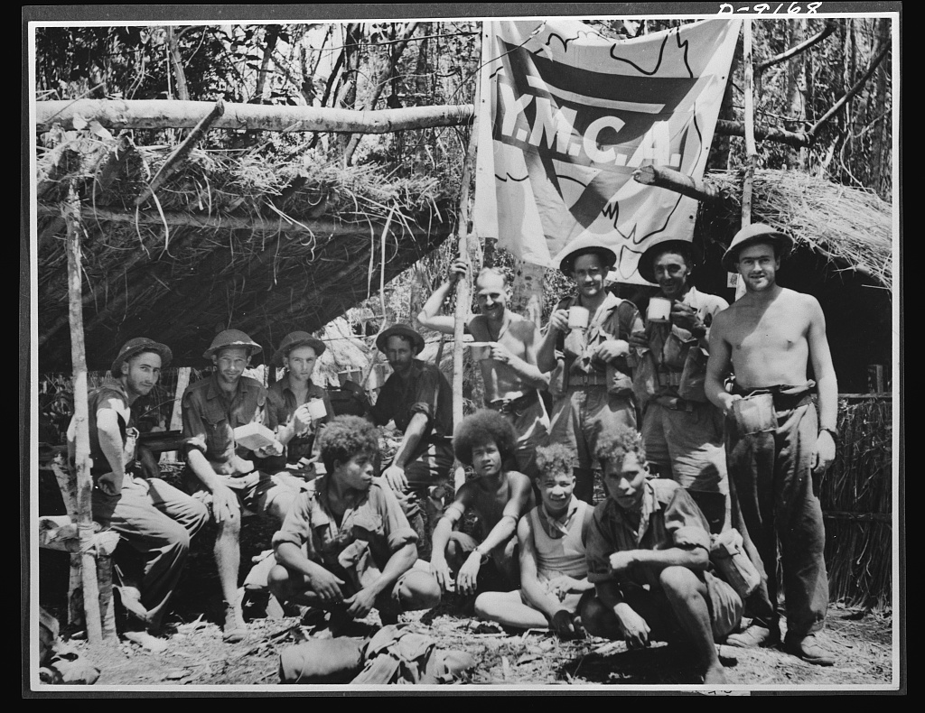 Natives aid Allied drive in New Guinea jungles. A short rest period in the New Guinea campaign. Natives and Allied troops pause at a YMCA (Young Men's Christian Association) unit in a forward area. These natives give invaluable aid to the Allied forces in transporting supplies and wounded soldiers through trackless jungles