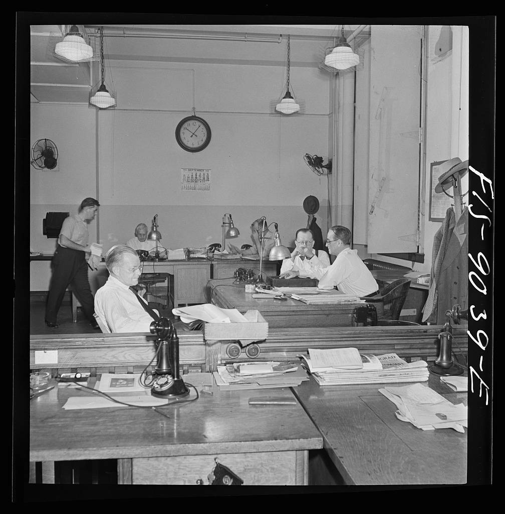 Newsroom of the New York Times newspaper. The "bull pen" where managing editors and others sit at one side. In foreground, assistant managing editor Rae. In background, managing editor James and night managing editor McCaw conferring