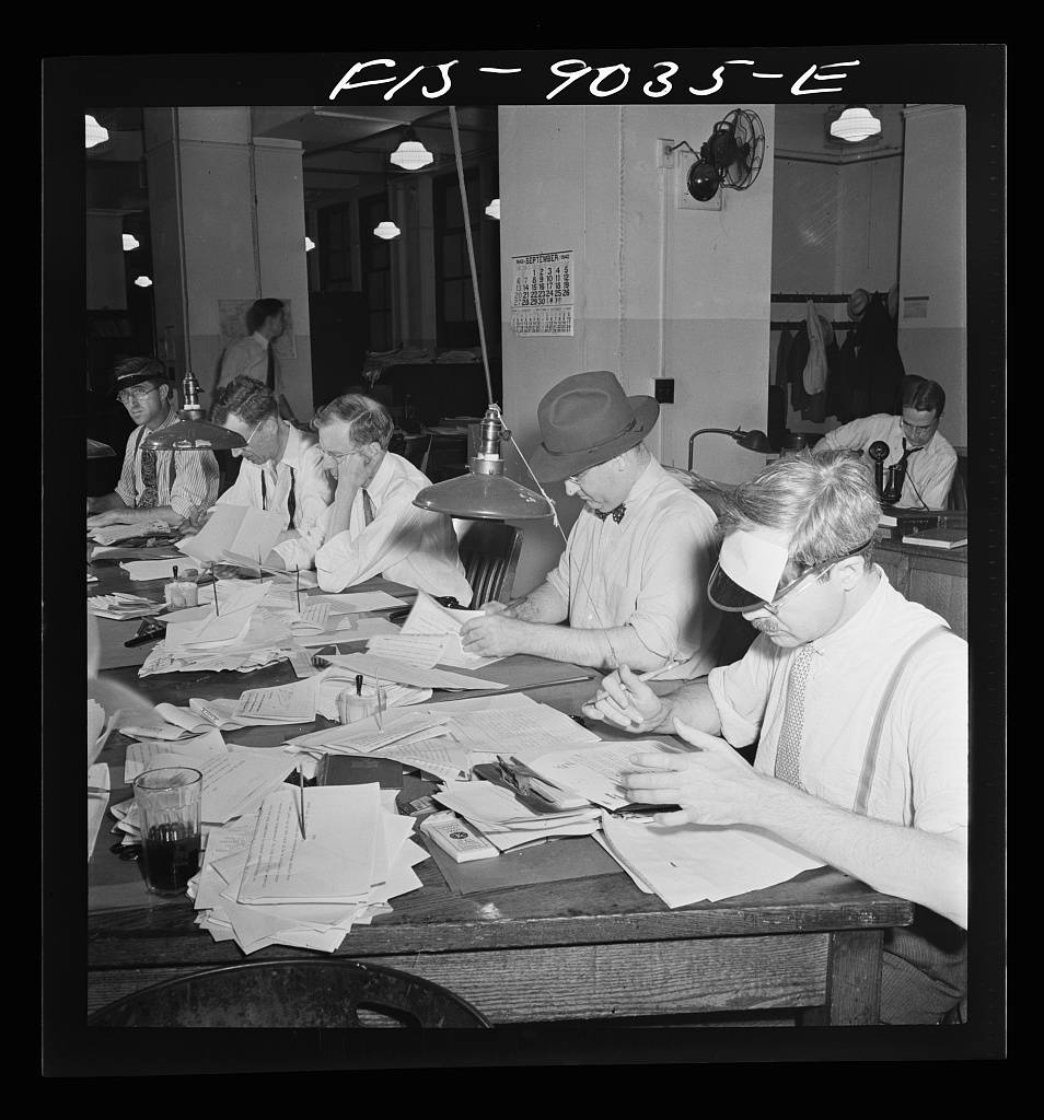 Newsroom of the New York Times newspaper. Copy readers at the telegraph desk which handles all dispatches from the U.S. outside New York City. Man wears hat because of draught