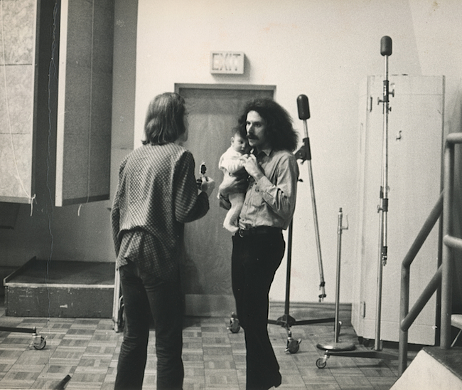 In a photo by Bob Cato, photographer Bob Seidemann is seen holding a band member’s baby while talking with Big Brother’s road manager, David Richards.