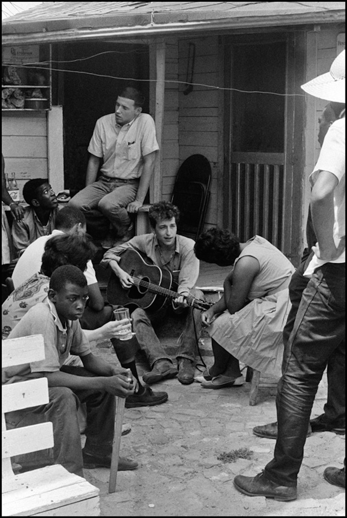 USA. Greenwood, Mississippi. 1962 After giving a concert in a cotton field with Pete Seeger and Theo Bikel, Bob Dylan plays behind the SNCC office. Bernice Reaon, one of the original Freedom Singers and later the leader of Sweet Honey in the Rock, listens. Mendy Samstein sits behind Dylan and talks to Willie Blue.