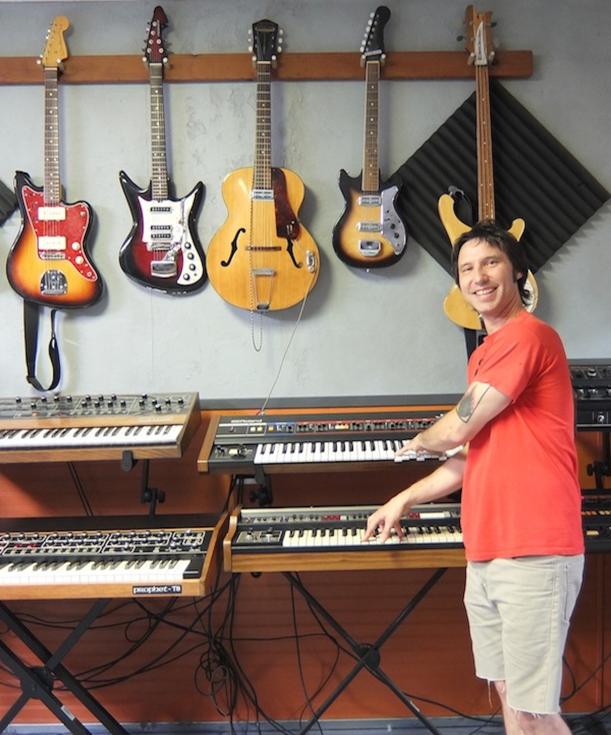 Lance Hill, goofing around with a pair or Roland synthesizers. The vintage guitars on the wall behind him are also used by artists who come to his studio to record