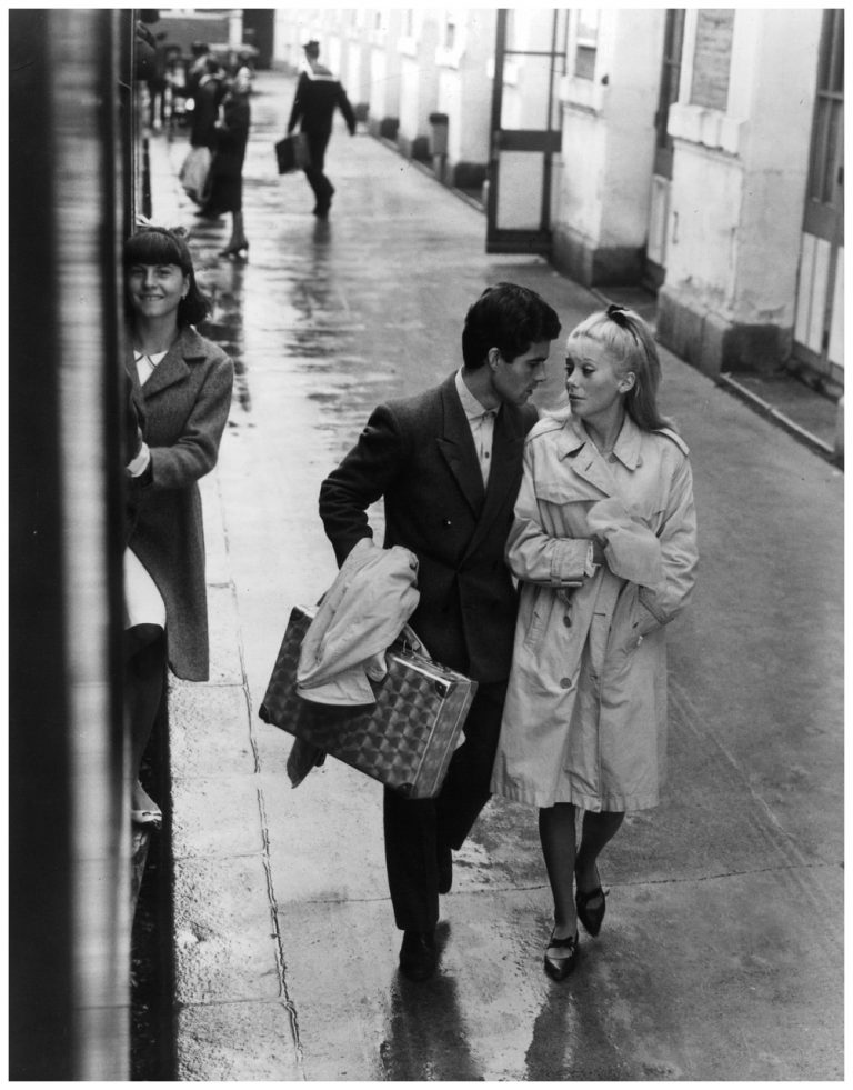 Nino Castelnuovo and Catherine Deneuve walk down a wet sidewalk in a ...