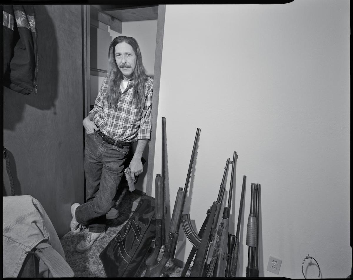 Photograph of a drug dealer standing in his home with his gun collection lining the wall.