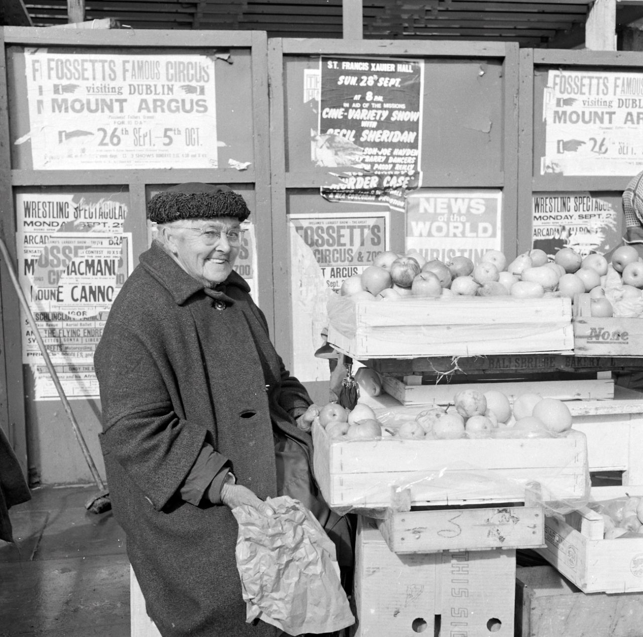 Ireland 1960s, Fruit seller with a very cheeky grin. We originally thought this photo was taken outside Heuston Station (previously Kingsbridge), Dublin, but see comments below showing it was taken alongside the hoarding around the site of the blown up Nelson's Pillar on O'Connell Street. It was definitely taken on the day of a G.A.A.match - perhaps the All-Ireland Final, given the dating evidence from the posters in the background. Anyone know this lady's name? Photographer: Elinor Wiltshire Date: September 1969