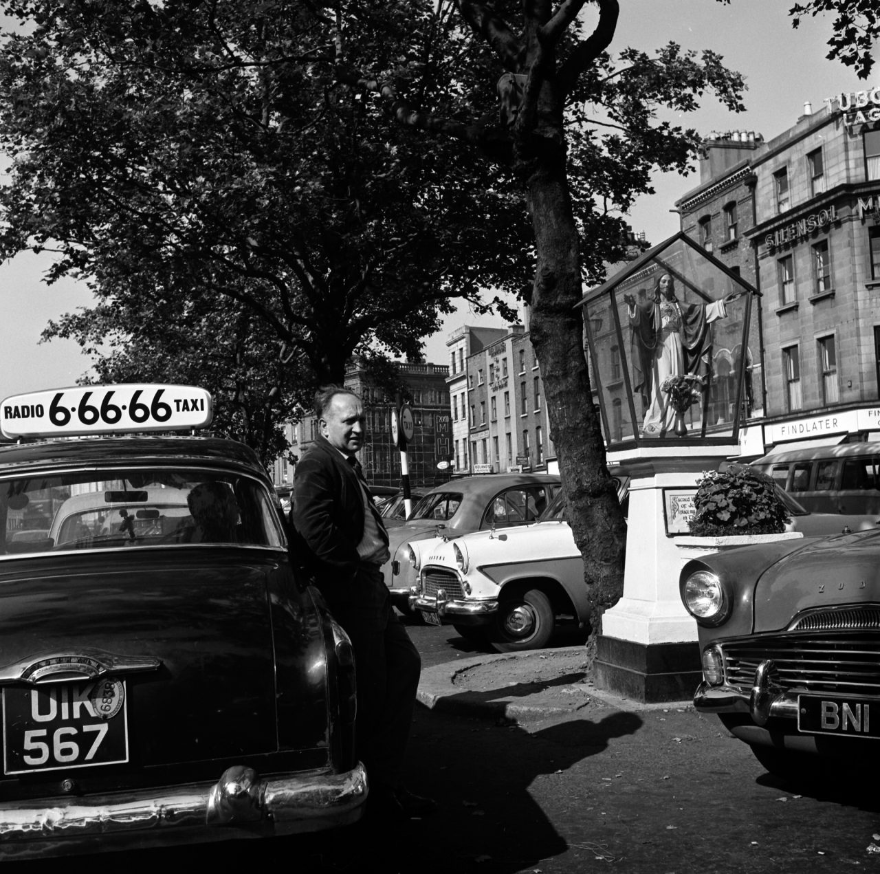 Ireland 1960s, Taxi driver waiting for a fare (and chatting to another driver leaning on his car?) at the rank in Dublin's O'Connell Street beside the Sacred Heart Shrine. Photographer: Elinor Wiltshire Date: Summer 1964