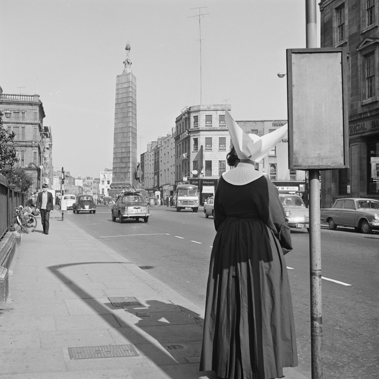 Ireland 1960s, A nun with a most remarkable (and slightly dangerous-looking) veil/head dress waiting for a bus outside the Rotunda Hospital on Parnell Street, Dublin. Cannot work out whether a clump of her own hair (unlikely) has come loose, or whether she is masking a person in front of her... With help from people over on our NLI Facebook page, it turns out this nun was a member of the Daughters of Charity (of St. Vincent de Paul). The distinctive head dress is called a cornette, and led to this order being known as the Butterfly Nuns. The Daughters of Charity abandoned the cornette on 20 September 1964, so unless this nun was ultra-traditional and carried on wearing it regardless, then this photo must have been taken before 20 September. This shot also allows us to view the Parnell Monument at the end of O'Connell Street from yet another angle. Photographer: Elinor Wiltshire Date: 1964