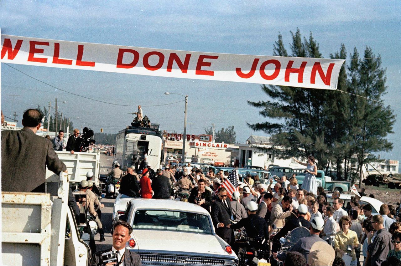 Astronaut John Glenn, left, sitting on back of car, is greeted by admirers as his car passes under a "Well Done John" banner in Cocoa Beach, Fla., . Glenn's wife Annie, center, dressed in red, and Vice President Lyndon Johnson are in the car with him