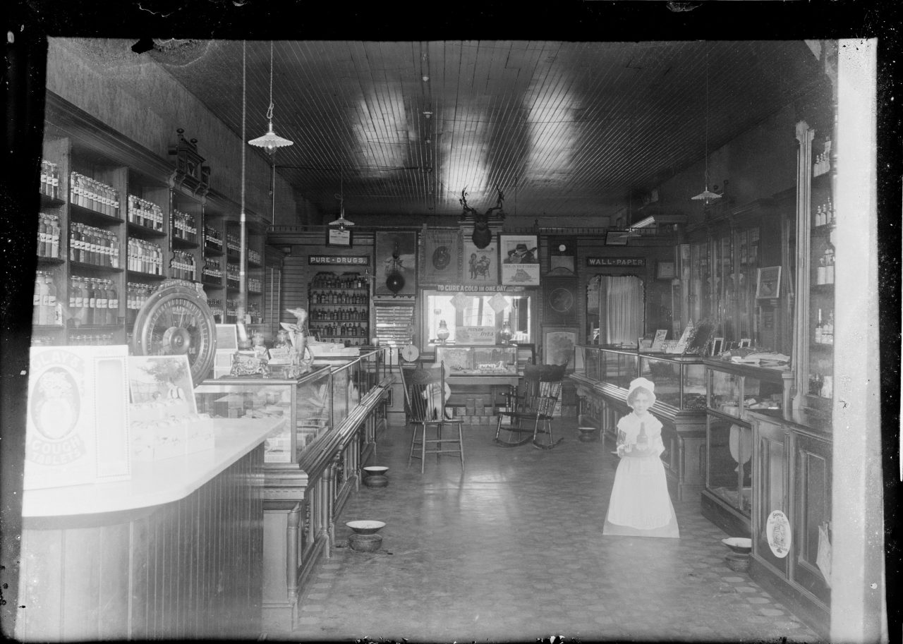 Shop with signs that read - Pure Drugs. To Cure A Cold In One Day. Wall PaperGeorge Silas Duntley Photographs 1899-1918