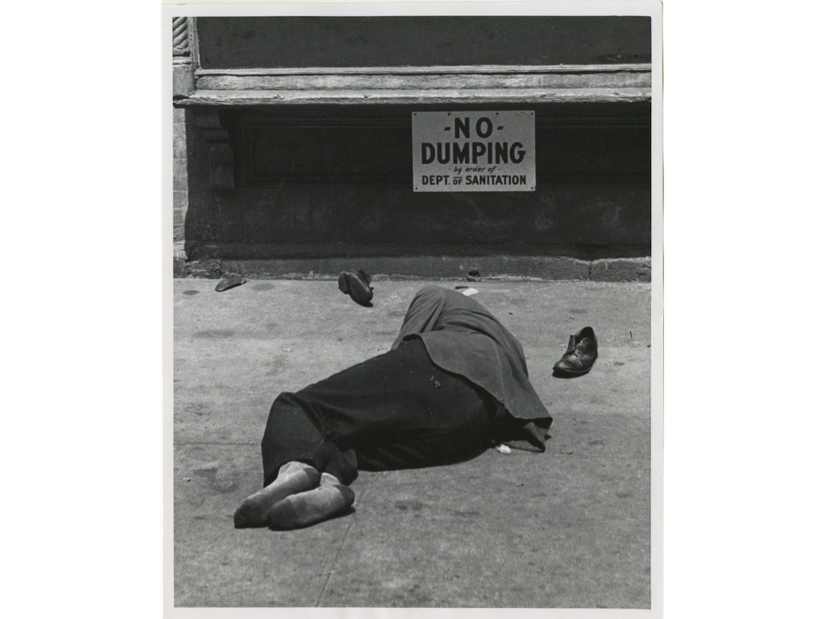 A man sleeps on the ground under a sign that reads NO DUMPING by order of Dept. of Sanitation on the Lower East Side.