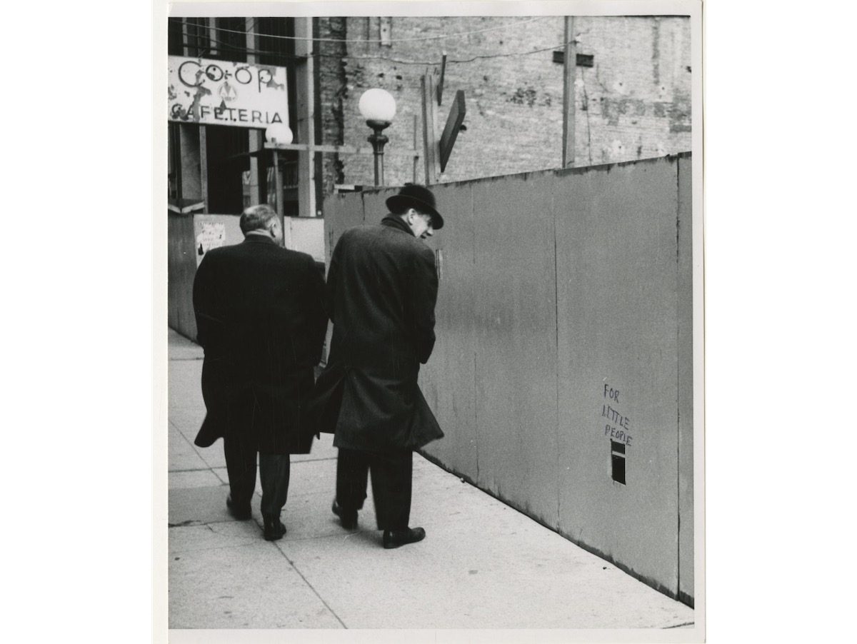Two men walk by a wooden wall with a rectangular cutout that says FOR LITTLE PEOPLE on the street in New York City. Inscription on verso- For Sidewalk Superintendents.