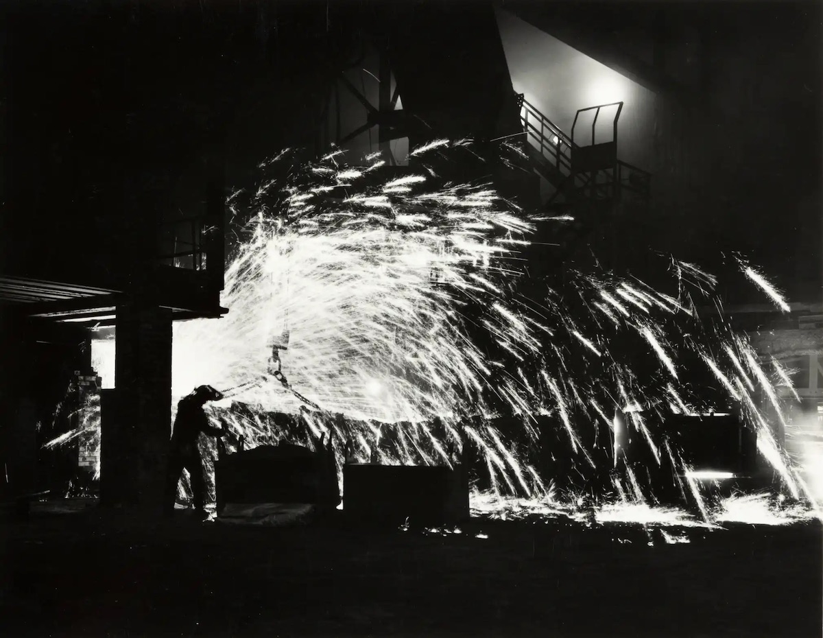 A front-side blast furnace worker settling a freshly cast furnace using vermiculite to calm the flow of molten iron from the furnace tap hole, 1975