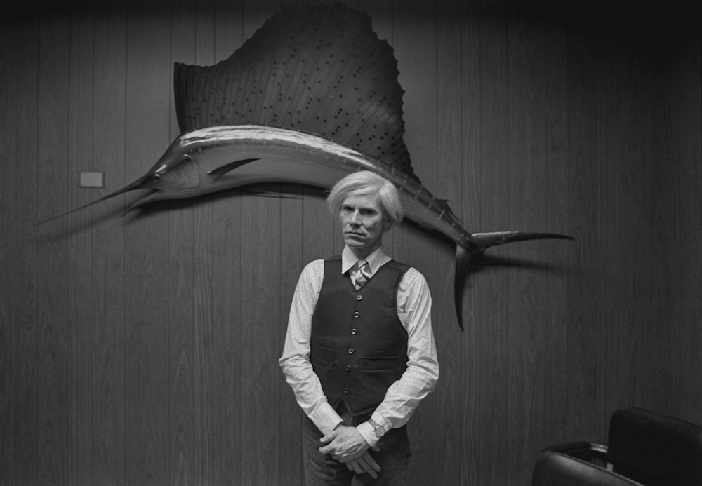 Andy Warhol posing at the New York City midtown store where he purchased a bullet proof vest, (1981).