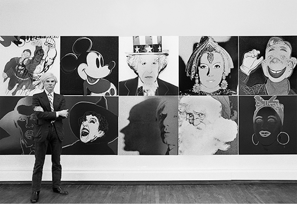 Andy Warhol standing in front of the installation of the Myths at the Robert Feldman Gallery in New York City (1981).