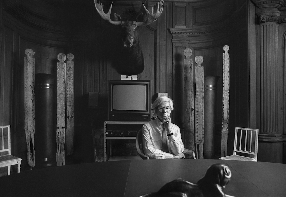 Andy seated in the conference room at the Factory with totems, VCR Television, and moose head taxidermy mounted on the wall (1981